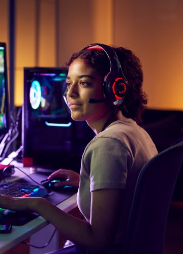 Teenage Girl Wearing Headset Gaming At Home Using Dual Computer Screens
